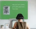 In a room with green plants and white walls, there's a green glass whiteboard hanging up. And there's a girl really getting down to work.